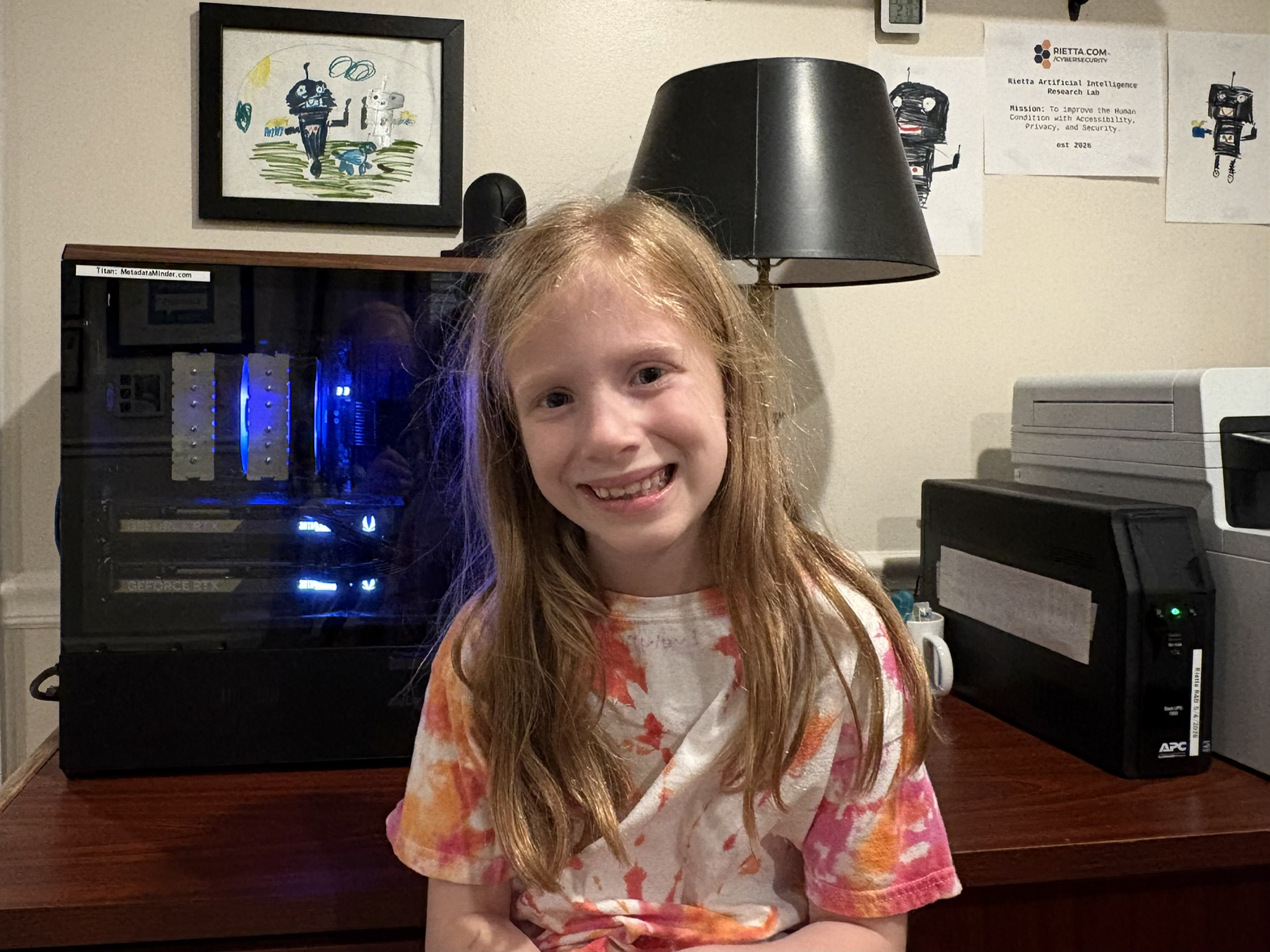The AMD 9900x + Dual RTX 5070 GPU server, Titan, a framed drawing of 'Robie, Graphie, and Tag', the Rietta AI Research Lab's mascots. My daughter Evelyn is smiling in the foreground, very proud of her artwork.