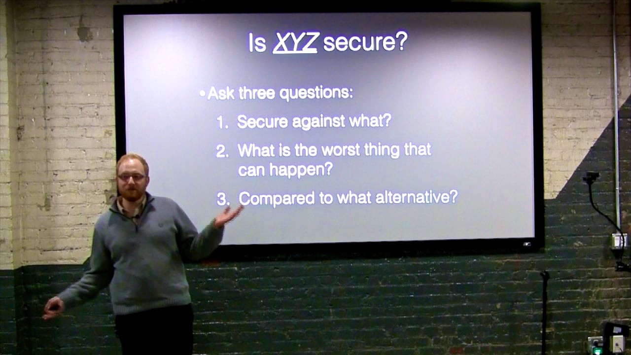 Frank Rietta standing beside a projector screen reading "Is XYZ secure? Ask three questions: secure against what, what's the worst thing that can happen, compared to what alternative."