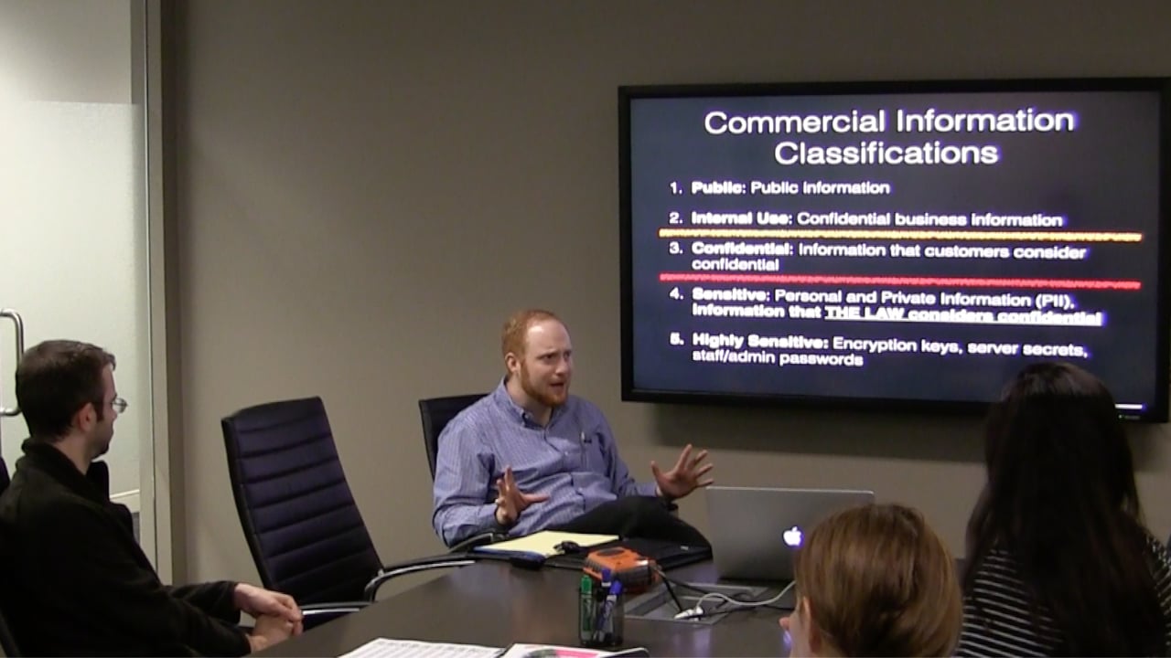 Frank Rietta presenting to colleagues around a conference table, with a screen behind him showing a "Commercial Information Classifications" slide listing five sensitivity levels.